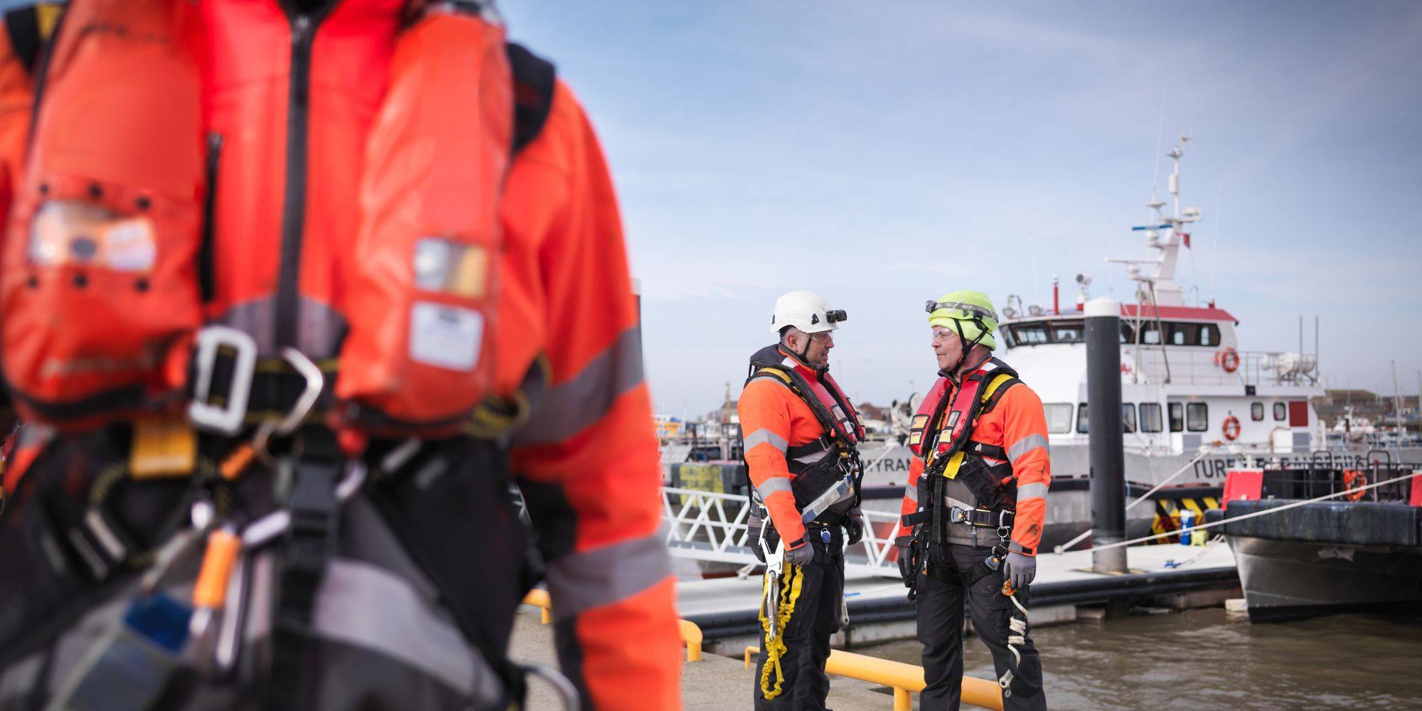 Two offshore safety workers in orange gear talking on a dock, with vessels docked behind them