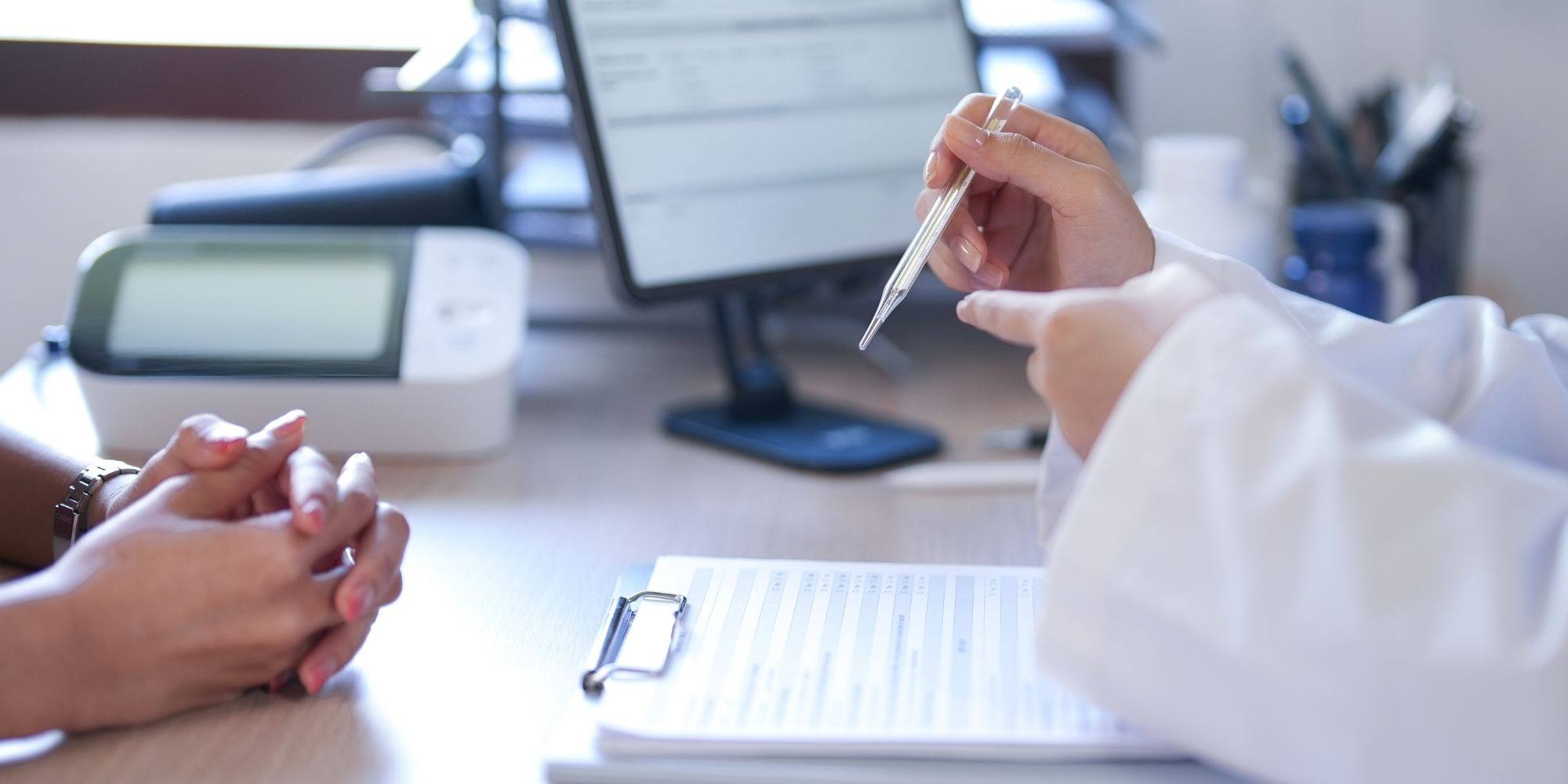 A clinician holding a thermometer with a patient's clasped hands nearby during an in-person consultation